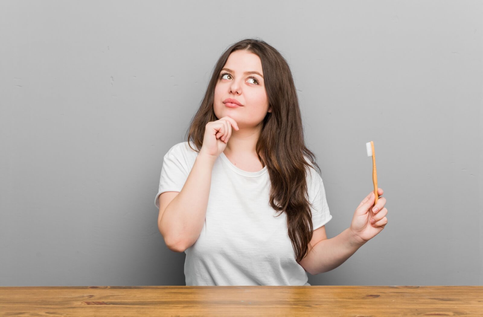 Person holding a yellow toothbrush against a grey wall wondering about dental hygiene practices.