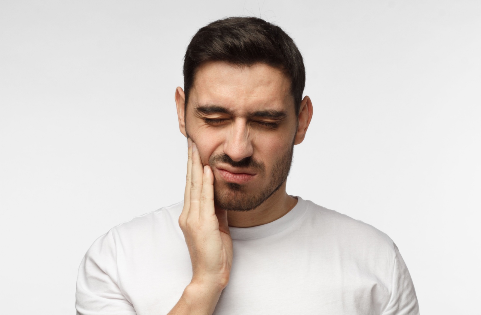 Person in a white t-shirt holding their cheek and wincing in pain, indicating a possible toothache from a cavity.
