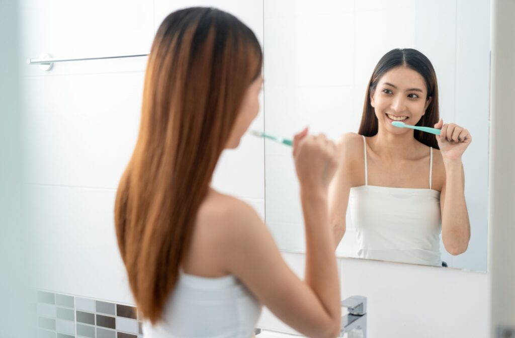 Person smiling while brushing their teeth in front of a bathroom mirror, wearing a white tank top.
