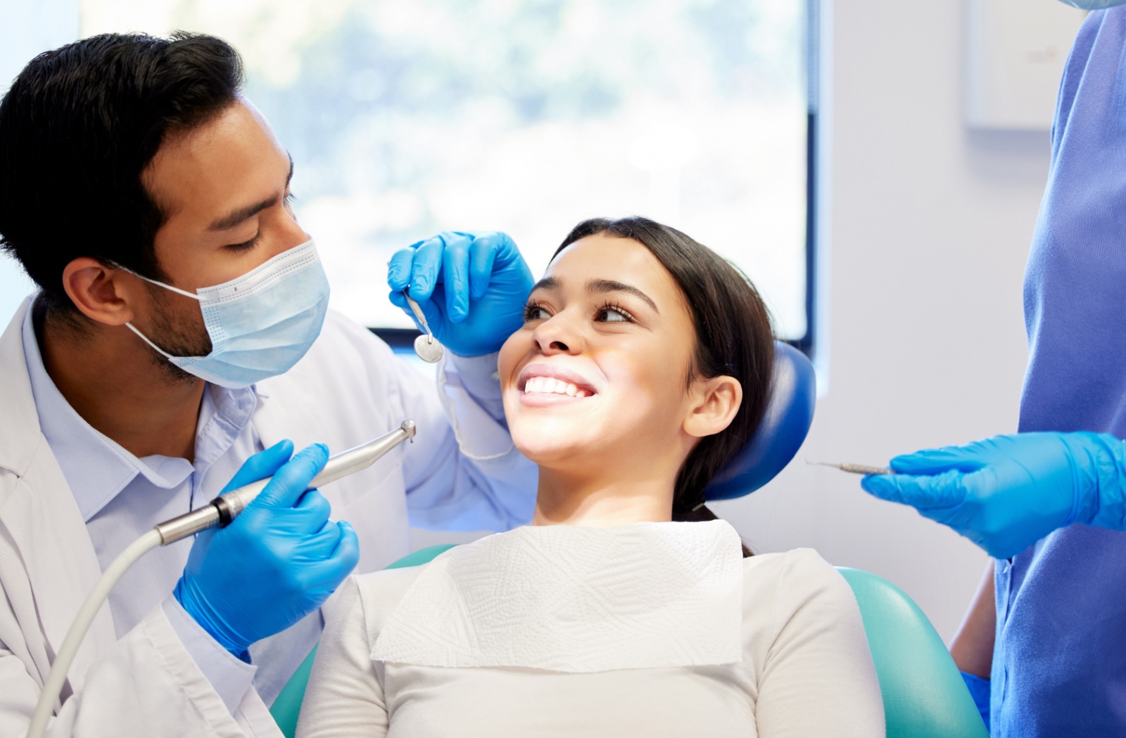 A person sits in a dental chair and smiles while a dentist wearing a mask and gloves prepares dental tools during an exam.