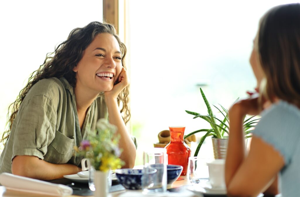 A couple friends sitting across the table from each other, laughing, enjoying the foods they love after a completed dental implant procedure.